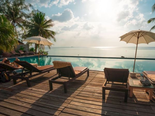 The image shows a serene poolside scene with lounge chairs, umbrellas, and a view of the ocean under a partly cloudy sky.
