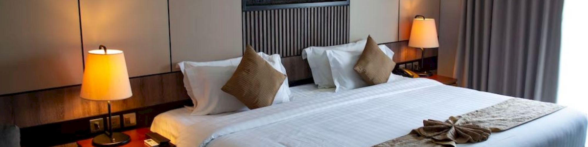 The image shows a neatly arranged hotel room with a large bed, stylish lamps, and decorative pillows, creating a cozy and inviting atmosphere.