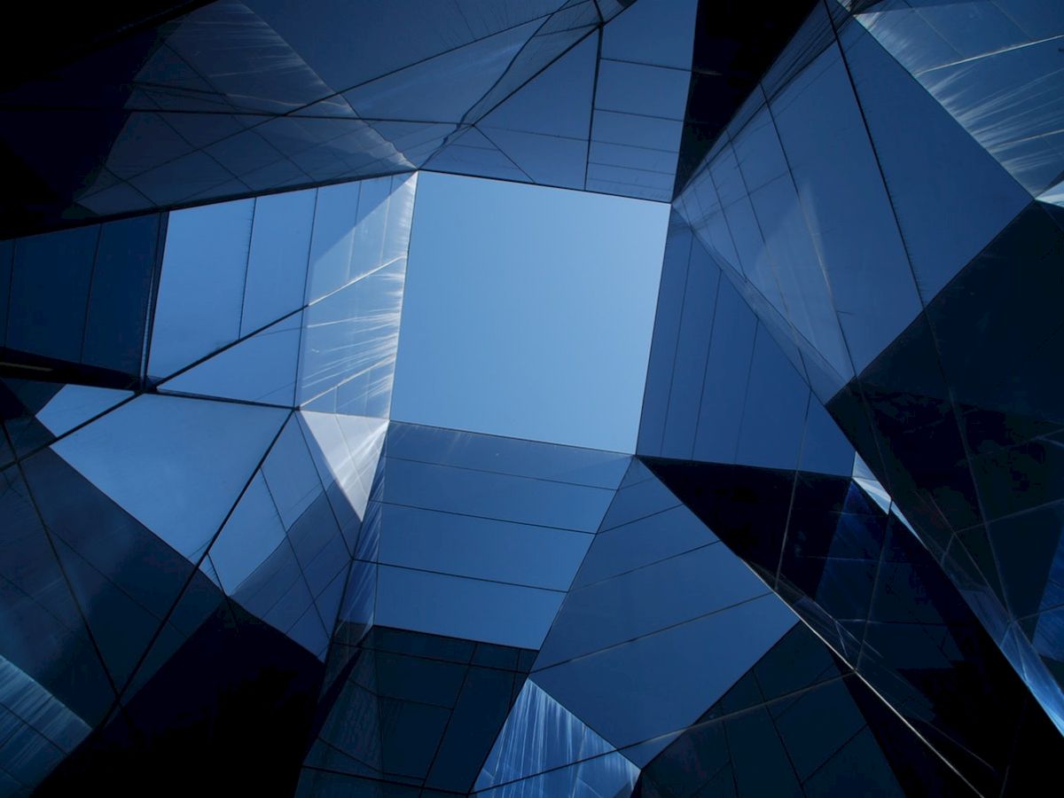 The image shows an abstract view looking up through geometric, mirrored structures with a clear blue sky visible at the center.