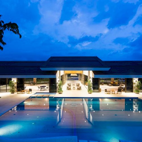 A luxurious modern building with a large, illuminated swimming pool at night, under a vibrant blue sky and lush surroundings.