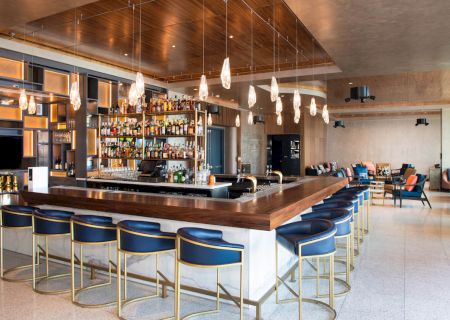 A modern bar with a wooden counter, blue chairs, hanging lights, and a stocked shelf. The warm ambiance suggests an inviting venue.