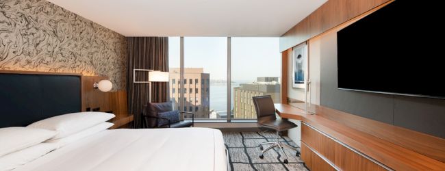 A modern hotel room with a large bed, TV, desk, chair, and window view of the cityscape.