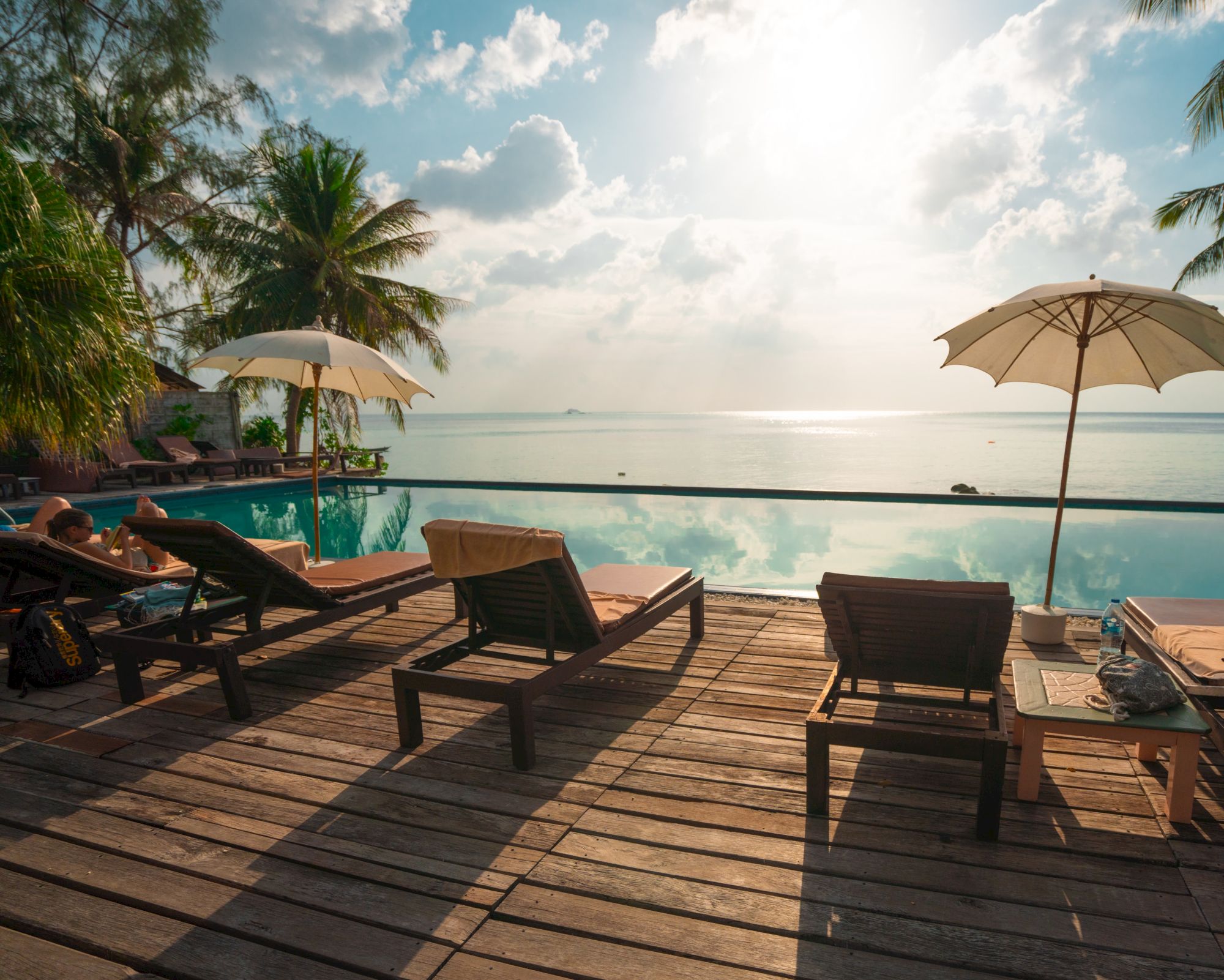 A tropical poolside with lounge chairs, umbrellas, palm trees, and a view of the ocean under a bright sky.