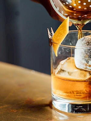 A cocktail is being poured into a glass with ice and a citrus peel garnish on a wooden surface.