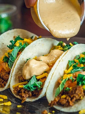 Three tacos filled with ground meat, cheese, and greens are on a plate; sauce is being poured over the middle one.