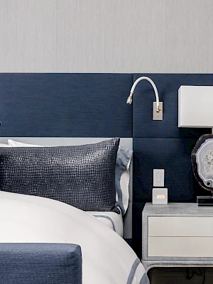 A modern bedroom features a blue and gray headboard, white bed linens, a nightstand with a lamp, and a decorative pillow.