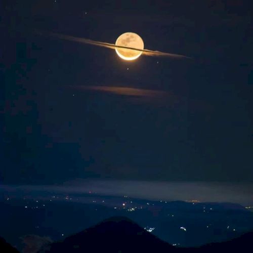 The image shows a full moon with a thin cloud crossing it, resembling Saturn's rings, over a landscape with distant lights below.