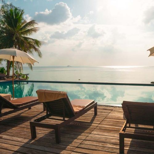 A serene beach scene with deck chairs, umbrellas, and palm trees by a pool, overlooking a calm sea under a partly cloudy sky.