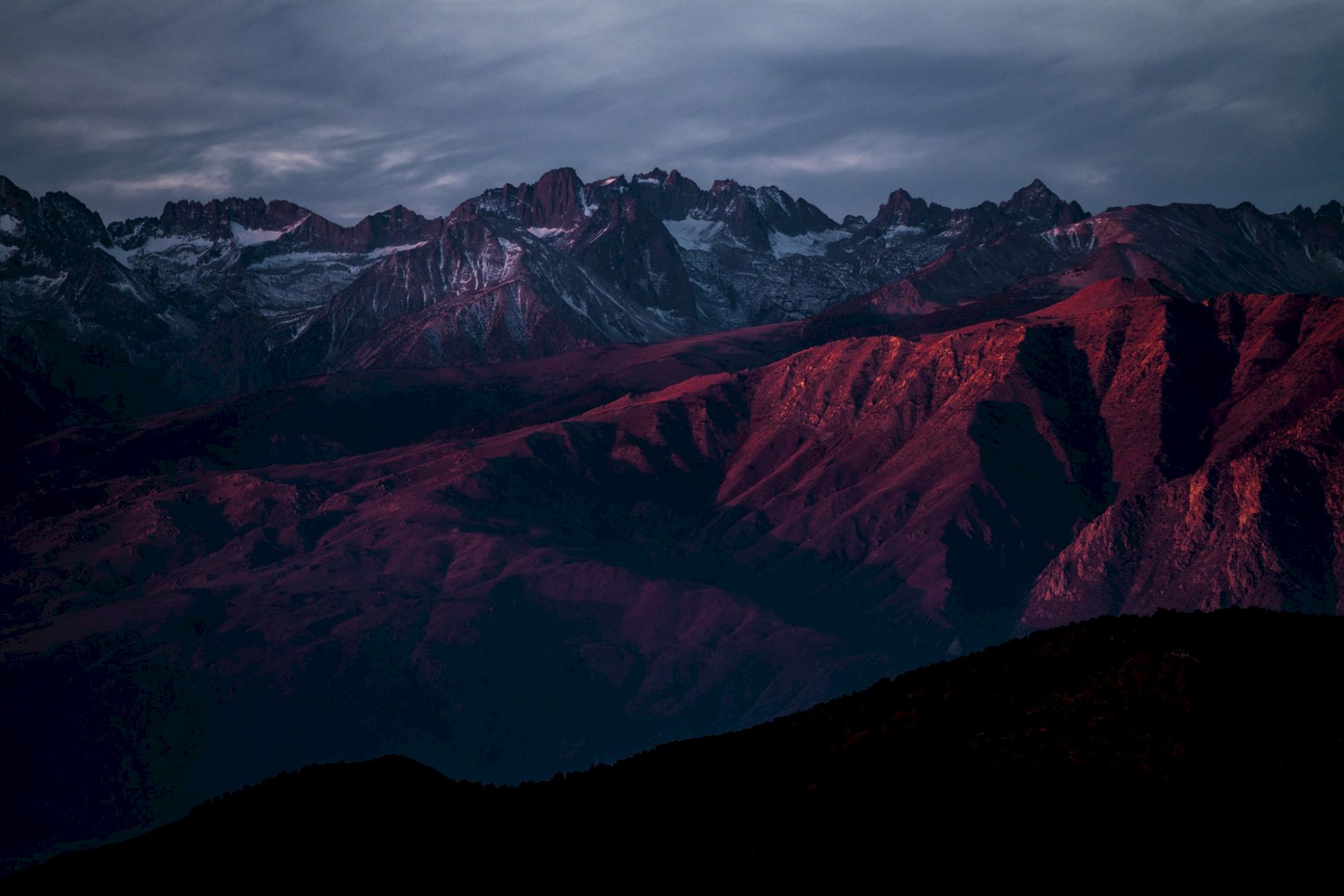 A mountain range at sunset, with peaks bathed in a reddish glow under a cloudy sky.