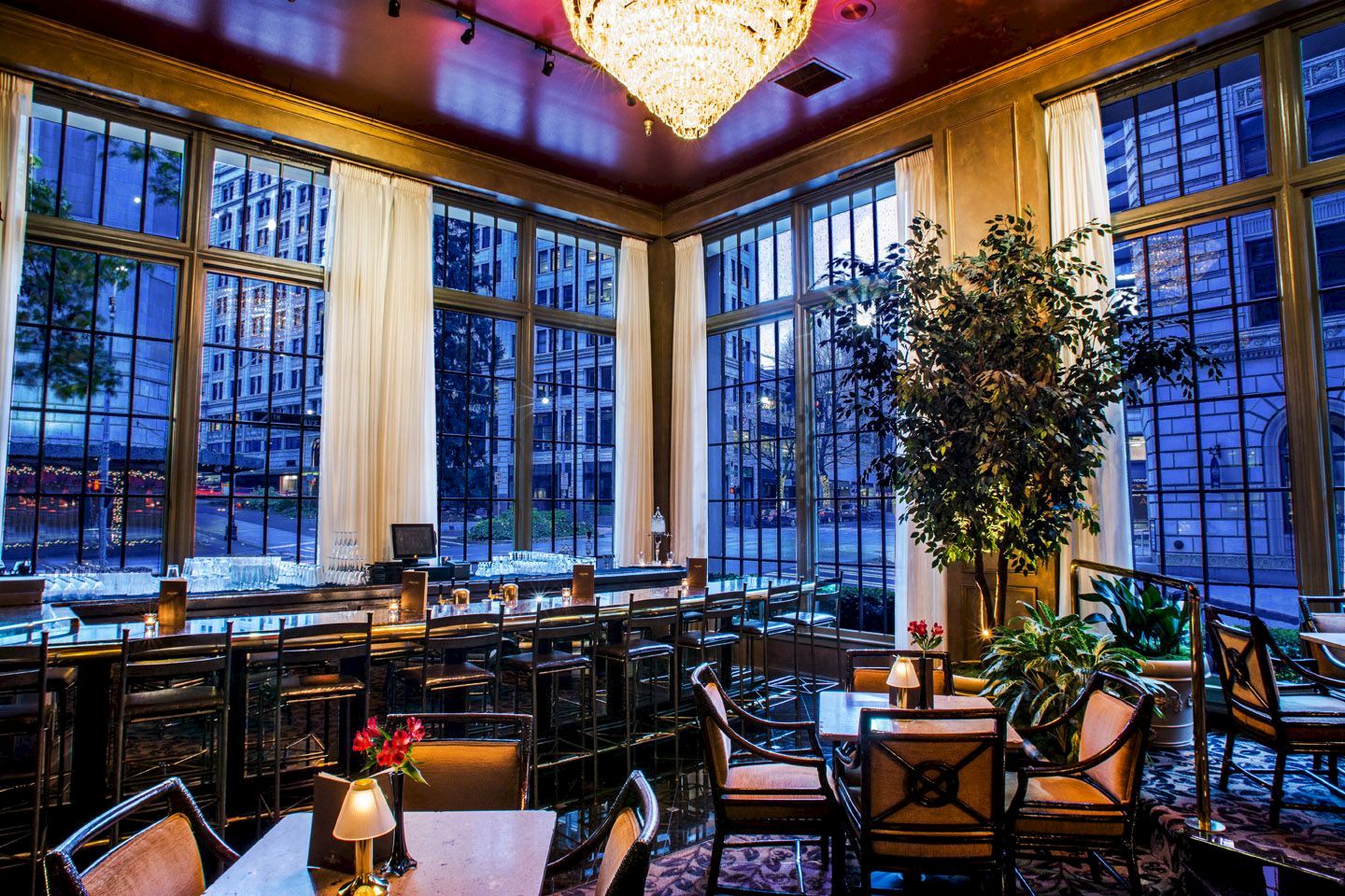 A chic restaurant interior with large windows, elegant furniture, a chandelier, and vibrant city views through the glass.