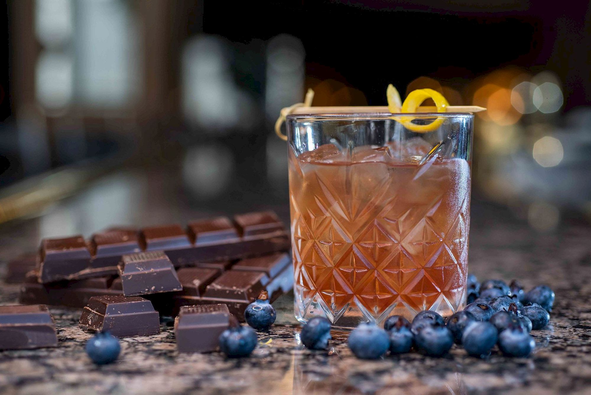 A beverage with lemon garnish, surrounded by chocolate and blueberries, is placed on a reflective surface with blurred lights in the background.