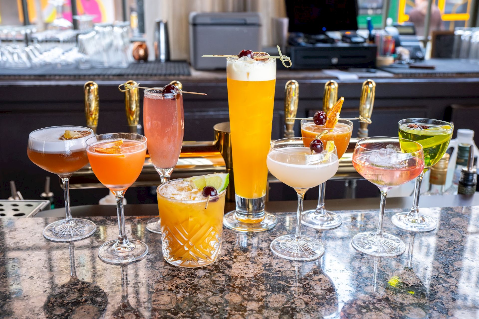 An array of colorful cocktails in various glasses is displayed on a bar counter, with a row of beer taps in the background.