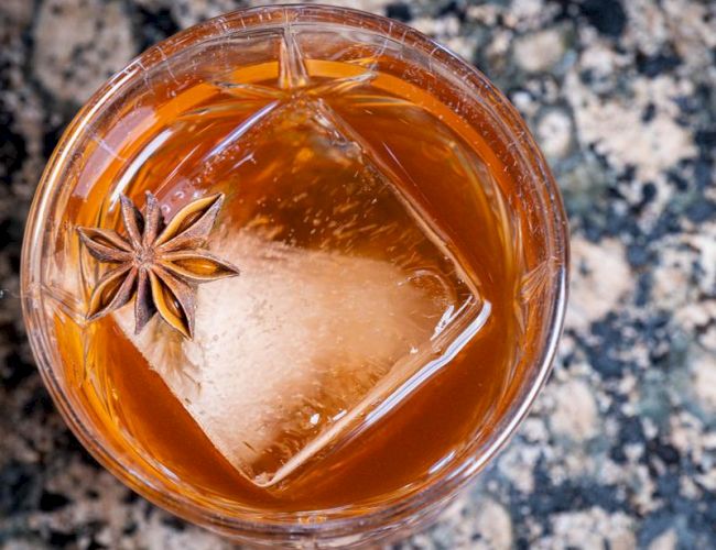 A glass with an amber liquid containing a large ice cube and a star anise on top, set on a speckled countertop.