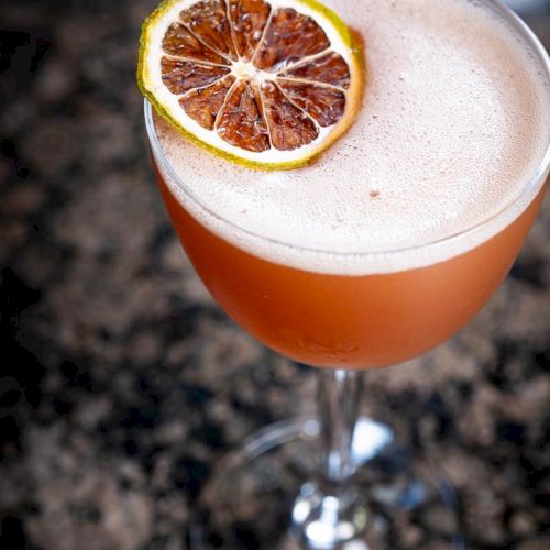 A frothy cocktail in a glass, garnished with a slice of dried citrus, on a dark, speckled surface.