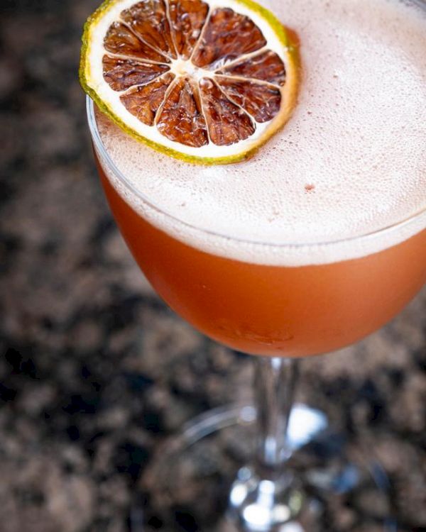 A frothy cocktail in a glass, garnished with a slice of dried citrus, on a dark, speckled surface.