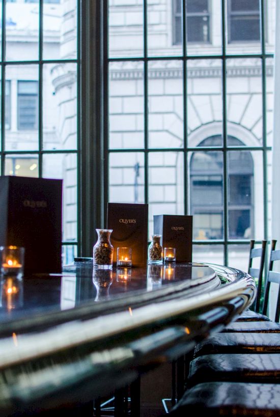 A stylish, dimly lit bar or restaurant with a curved counter, candles, and rows of chairs, view of a large window and city buildings beyond.