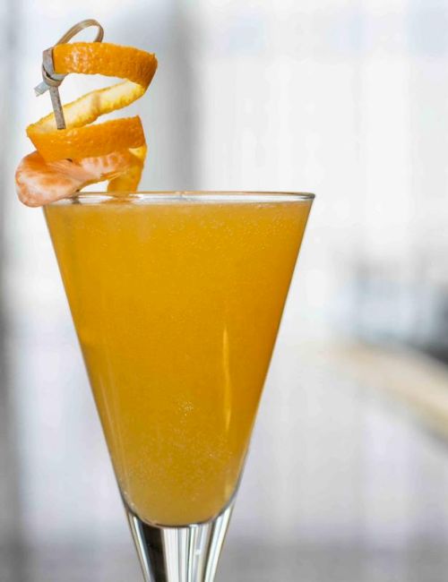 A tall glass with an orange cocktail, garnished with a spiral peel and a cherry, sits on a blurred background.