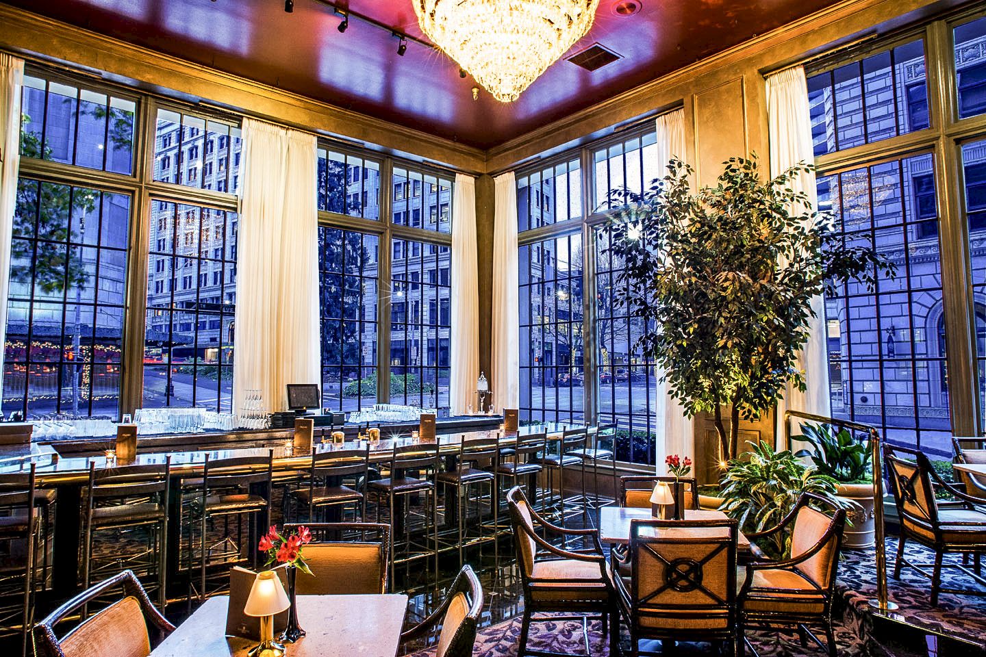 A stylish, sunlit dining area with tall windows, plants, and cozy seating by a bar&mdash;elegant, urban, and inviting.