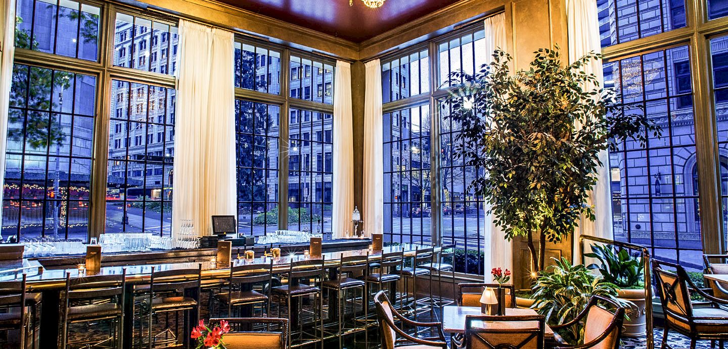 A stylish, sunlit dining area with tall windows, plants, and cozy seating by a bar&mdash;elegant, urban, and inviting.