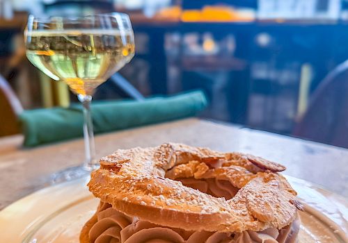 A fancy pastry with cream swirls on a cookie shell, dusted with powdered sugar, served on a plate with a glass of white wine in a bright restaurant.