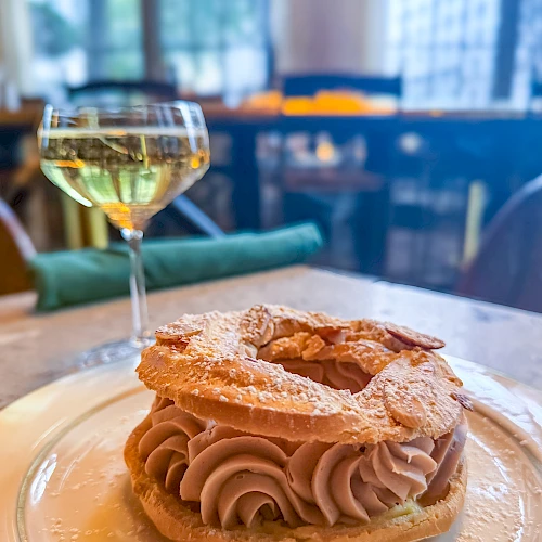 A fancy pastry with cream swirls on a cookie shell, dusted with powdered sugar, served on a plate with a glass of white wine in a bright restaurant.