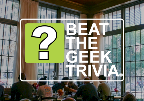 A conference room with people seated for a meal while a &ldquo;Beat the Geek Trivia&rdquo; banner overlays the scene beneath a chandelier.