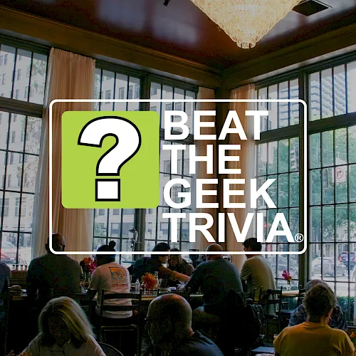 A conference room with people seated for a meal while a “Beat the Geek Trivia” banner overlays the scene beneath a chandelier.