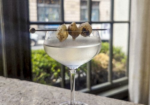 A coupe glass with a clear cocktail, garnished with three lime-green olives on a skewer, set on a bar counter by a window.