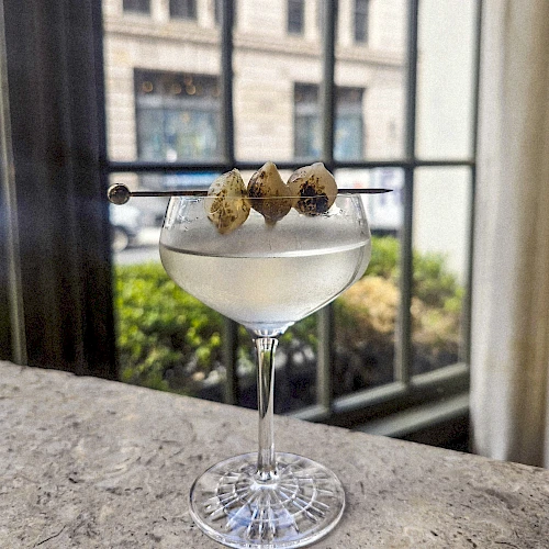 A coupe glass with a clear cocktail, garnished with three lime-green olives on a skewer, set on a bar counter by a window.