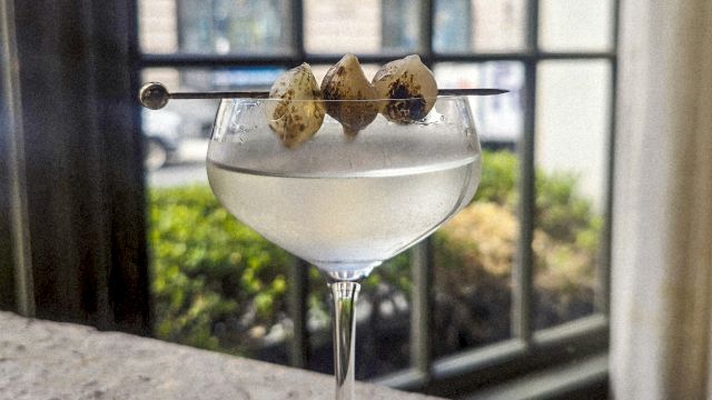 A coupe glass with a clear cocktail, garnished with three lime-green olives on a skewer, set on a bar counter by a window.