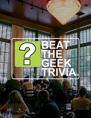 A restaurant meeting scene with a sign reading &ldquo;BEAT THE GEEK,&rdquo; featuring a big green question mark, in front of diners.