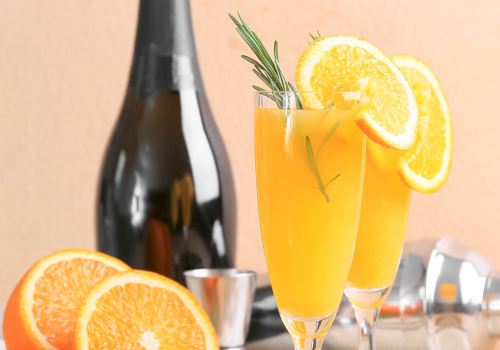 A bottle of sparkling wine with two orange cocktails garnished with orange slices and rosemary, surrounded by fresh orange halves.