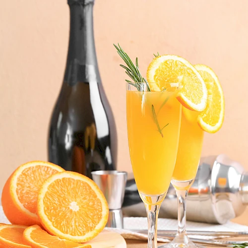 A bottle of sparkling wine with two orange cocktails garnished with orange slices and rosemary, surrounded by fresh orange halves.