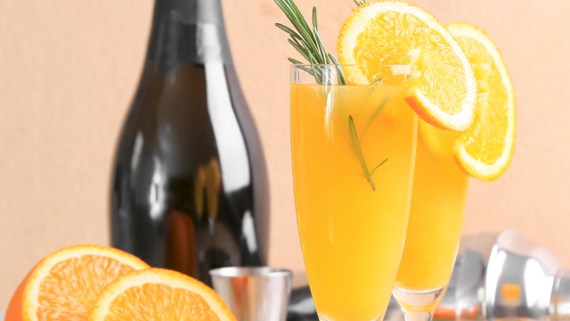 A bottle of sparkling wine with two orange cocktails garnished with orange slices and rosemary, surrounded by fresh orange halves.