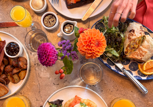 Brunch spread with stacked French toast/waffles, sunny eggs, salad and pastries, juice and coffee, plus fresh flowers on the table.