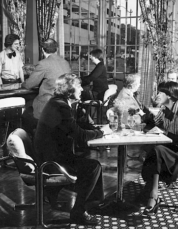 A group of people dine and chat in a classic restaurant booth, with a waiter nearby and drinks on the table.
