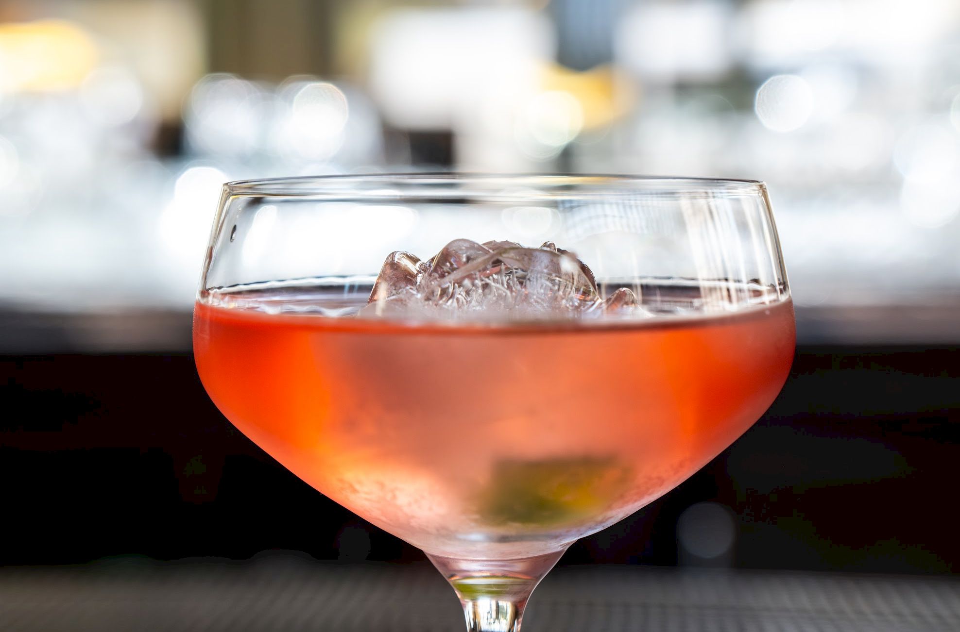 A stemmed glass filled with an orange drink and ice cubes, with a softly blurred background of lights.