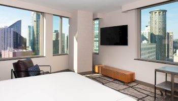 A modern hotel room with large windows showing a cityscape, featuring a bed, wall-mounted TV, armchair, and a stylish rug on the floor.