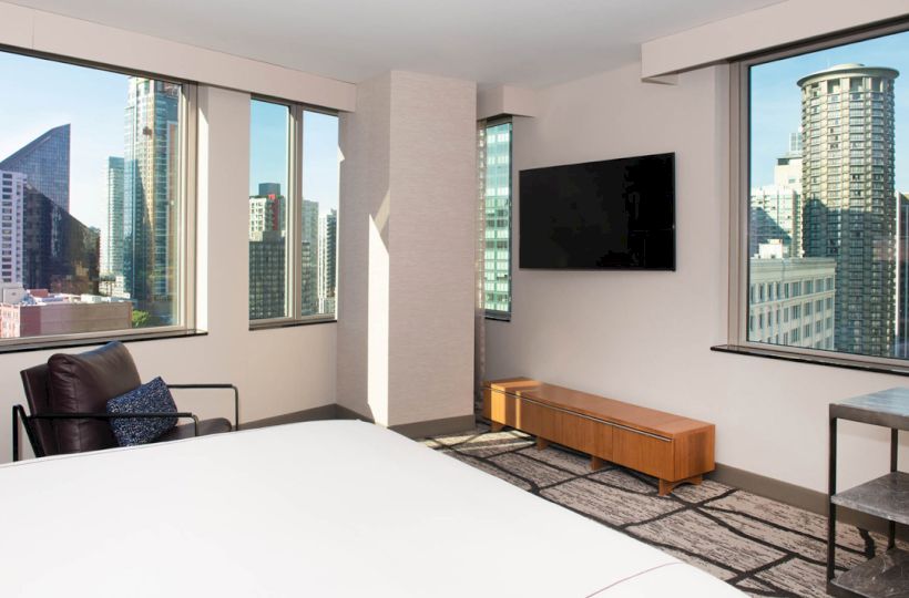 A modern hotel room with large windows showing a cityscape, featuring a bed, wall-mounted TV, armchair, and a stylish rug on the floor.