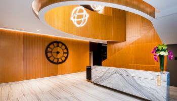 A modern lobby with wooden panels, a marble desk, a large clock, and spherical pendant lights, featuring flower arrangements on the desk.
