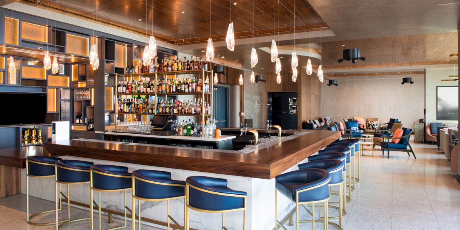 The image shows a stylish bar with a wooden counter, blue barstools, pendant lights, and a well-stocked shelf behind the counter.