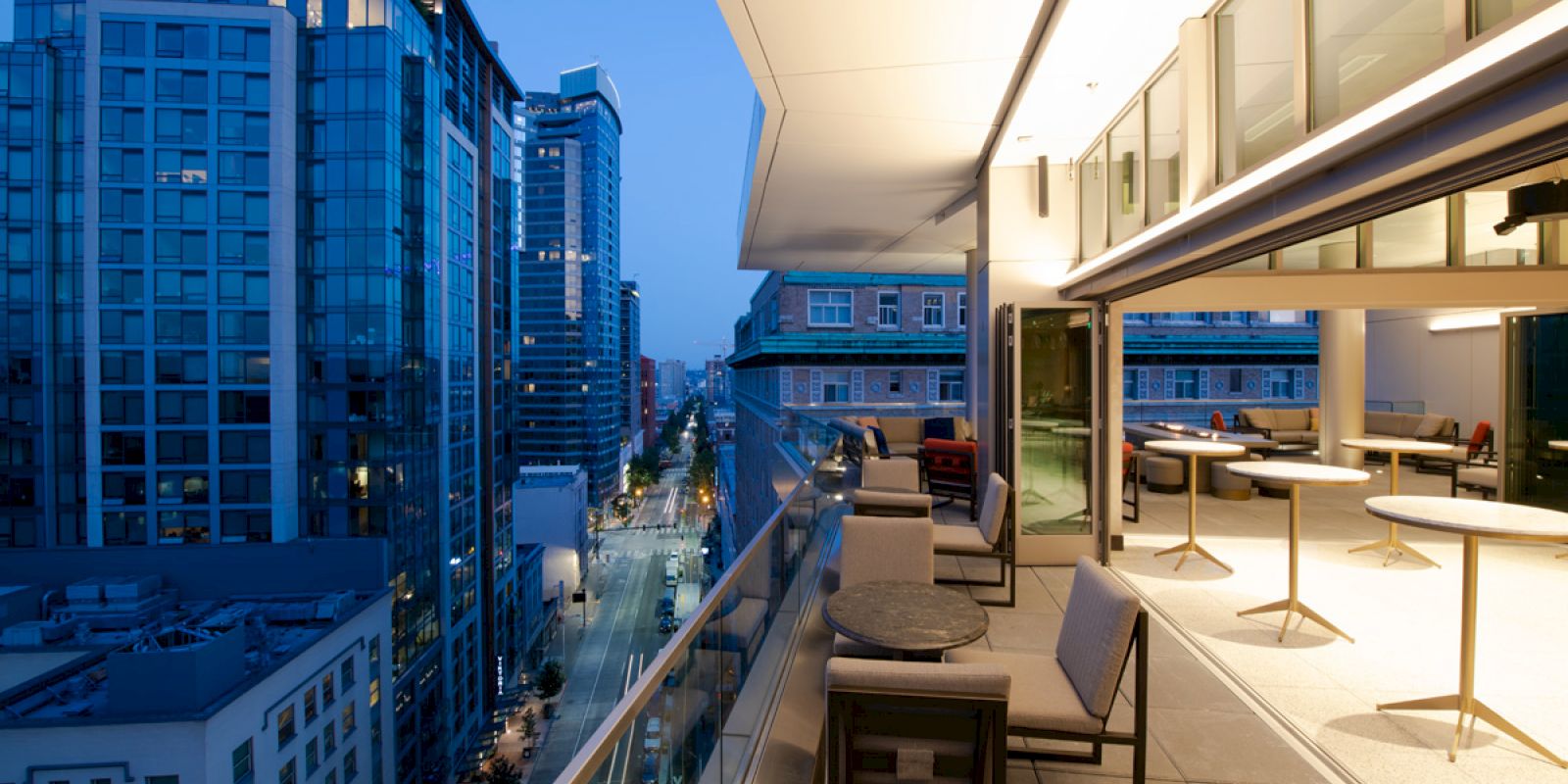 A rooftop terrace with seating overlooks a cityscape of modern buildings at twilight.