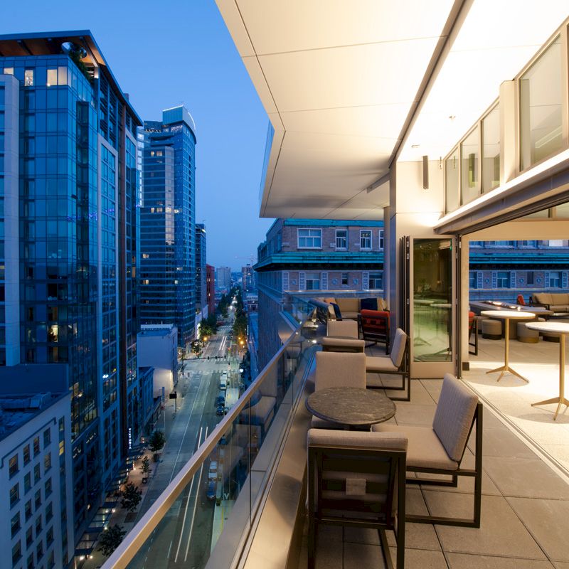 A modern rooftop terrace with seating overlooks a cityscape of tall buildings and a street below during early evening.