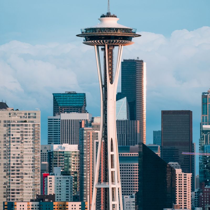 The image shows the Space Needle in Seattle, surrounded by various skyscrapers and buildings against a cloudy sky backdrop.
