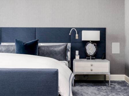A modern bedroom with a blue headboard, white bedding, a nightstand with a lamp, decor item, and a wall-mounted reading light.