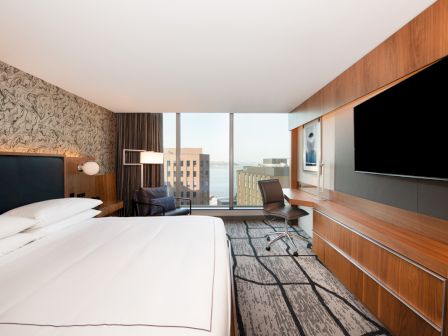 A modern hotel room with a large bed, wall-mounted TV, desk, and window with an urban view is depicted in the image.