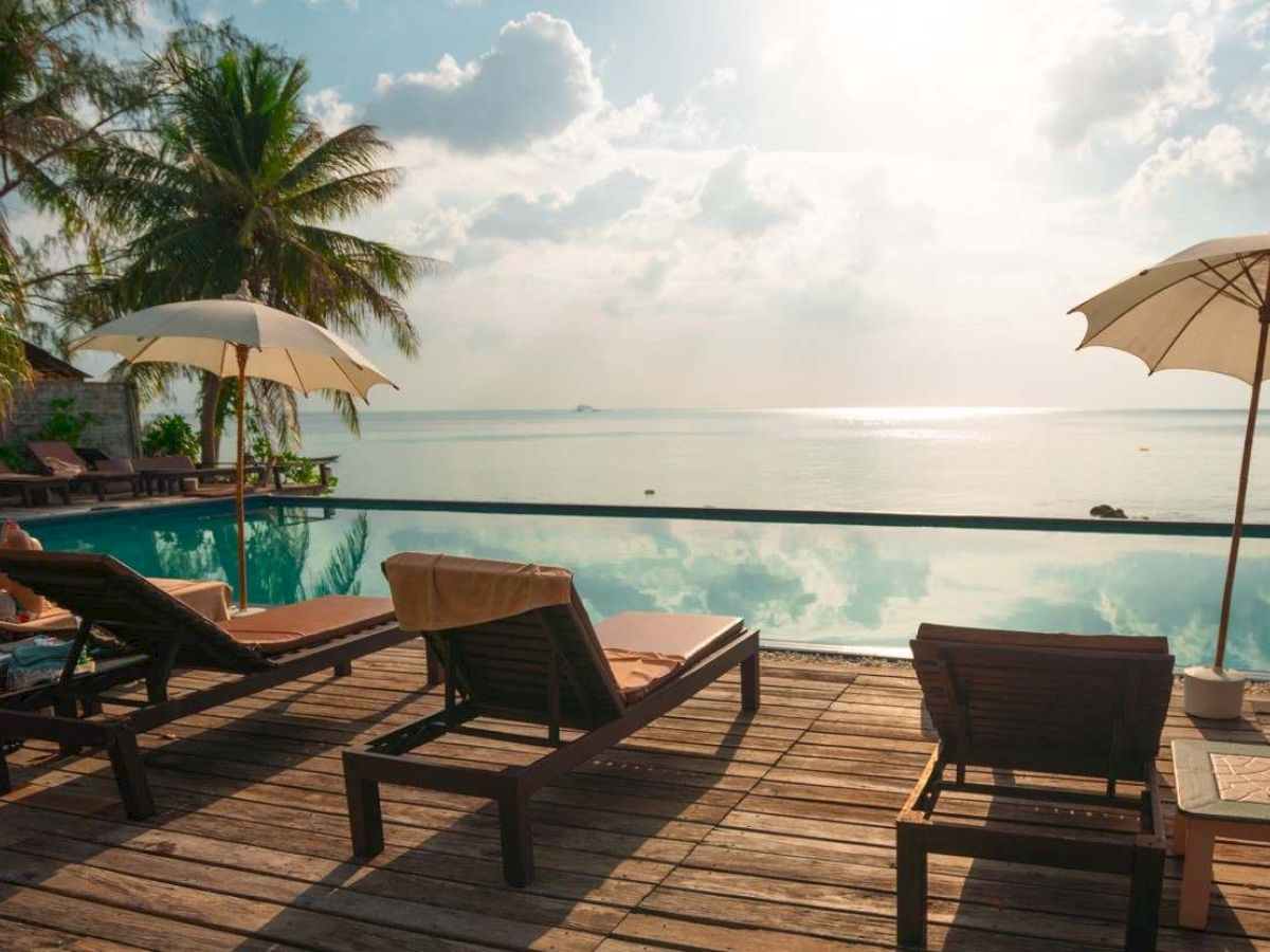 Poolside view with lounge chairs, umbrellas, and palm trees overlooking a calm sea and cloudy sky.