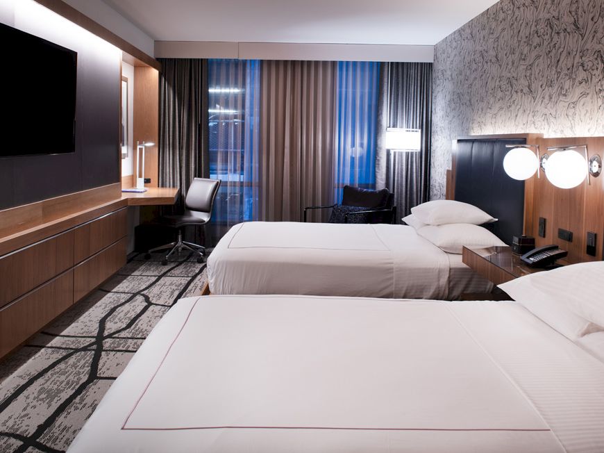 The image shows a hotel room with two beds, a wall-mounted TV, a desk, lamps, and curtains, combining modern and cozy elements.