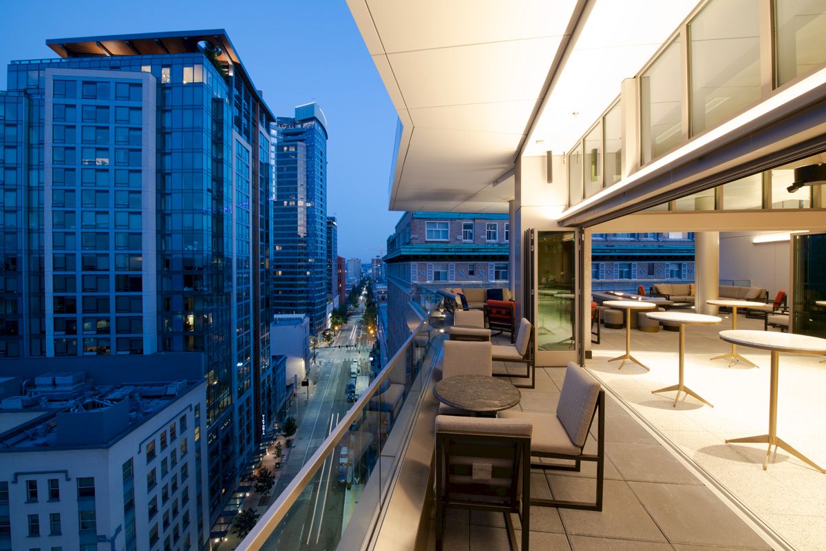 A modern rooftop terrace with city views, featuring seating areas and tables, overlooking a street lined with tall buildings at dusk.