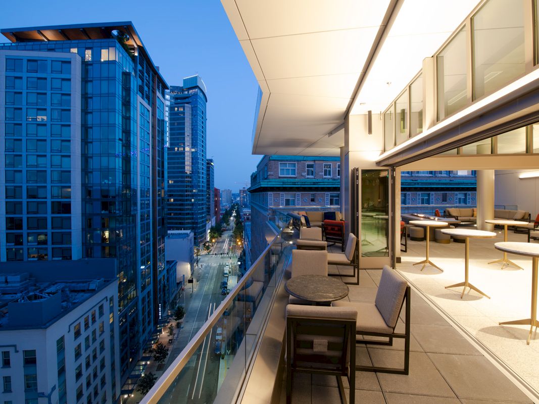 A modern rooftop terrace with city views, featuring seating areas and tables, overlooking a street lined with tall buildings at dusk.